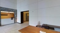 This single-floor space makes an immediate impression with its dramatic ceiling height and expansive large-format wall panels in a clean, neutral palette. The interplay of materials is a defining characteristic — smooth, crisp white paneling contrasts beautifully with a rugged natural stone accent wall, creating a dynamic tension between modern minimalism and organic texture. Light-toned tile flooring runs throughout, adding to the open, airy feel of the space. A warm wood reception or display surface anchors the main area, complemented by dark-toned cabinetry and tasteful metallic decorative accents, including sculptural leaf motifs that add an artistic touch. A recessed doorway leads to an adjacent corridor or secondary area, hinting at further functional spaces beyond. Subtle accent lighting highlights the stone wall and architectural transitions, adding warmth and depth to the overall composition. The combination of soaring vertical space, clean lines, natural stone, and carefully chosen furnishings gives this space a polished, professional atmosphere — well-suited for a reception area, lobby, or upscale commercial environment.