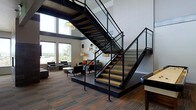 This two-story space is defined by its bold industrial-modern aesthetic, anchored by a dramatic open-tread staircase with a sleek black steel frame and glass panel railings. The staircase serves as the architectural centerpiece, connecting both floors while allowing sightlines to flow freely throughout the space. Double-height ceilings amplify the sense of volume and grandeur, while floor-to-ceiling windows flood the interior with abundant natural light and frame sweeping outdoor views of the surrounding landscape.

The main level features a lounge-style seating area positioned on a raised platform, furnished with contemporary chairs and sofas in neutral tones accented with pops of color. Warm-toned wood flooring and richly patterned carpet tiles ground the space, adding texture and depth. Accent lighting from standing lamps complements the natural light, creating a welcoming ambiance. A colorful wall-mounted art display adds a creative focal point to the interior. The overall atmosphere is sophisticated yet approachable, combining open, airy proportions with carefully curated furnishings that encourage both relaxation and social interaction.