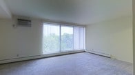 This single-floor space offers a clean, functional layout with a calm and understated aesthetic. The living area is a standout feature, highlighted by a large sliding glass door fitted with vertical blinds that allow for excellent light control. The expansive window wall floods the room with natural light while framing a peaceful view of surrounding greenery and trees, creating a tranquil indoor-outdoor connection. The living space is finished with wall-to-wall carpeting in a neutral tone, contributing to a warm and comfortable atmosphere. Baseboard heating runs along the perimeter of the room, providing an efficient and unobtrusive climate control solution. The walls are finished in a soft, neutral palette that complements the natural light and keeps the space feeling open and airy. The overall design is straightforward and practical, offering a versatile canvas suitable for a variety of furniture arrangements and personal styles. The single-floor layout ensures ease of movement throughout the space, making it accessible and convenient. With its abundant natural light, outdoor views, and functional design, this space provides a comfortable and inviting environment.