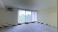 This single-floor space offers a clean, functional layout with a calm and understated aesthetic. The living area is a standout feature, highlighted by a large sliding glass door fitted with vertical blinds that allow for excellent light control. The expansive window wall floods the room with natural light while framing a peaceful view of surrounding greenery and trees, creating a tranquil indoor-outdoor connection. The living space is finished with wall-to-wall carpeting in a neutral tone, contributing to a warm and comfortable atmosphere. Baseboard heating runs along the perimeter of the room, providing an efficient and unobtrusive climate control solution. The walls are finished in a soft, neutral palette that complements the natural light and keeps the space feeling open and airy. The overall design is straightforward and practical, offering a versatile canvas suitable for a variety of furniture arrangements and personal styles. The single-floor layout ensures ease of movement throughout the space, making it accessible and convenient. With its abundant natural light, outdoor views, and functional design, this space provides a comfortable and inviting environment.