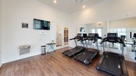 The fitness center at 900 Graces Reserve Circle in Kannapolis, NC is a well-equipped, single-floor community facility designed to support a comprehensive workout experience. The space features warm hardwood-style flooring throughout, crisp white walls, crown molding, and recessed lighting that create a clean, inviting atmosphere. Elevated ceilings with tray detailing add a sense of openness to the room.

Cardio equipment is prominently featured, with multiple treadmills and elliptical trainers positioned for easy use. A dedicated free weight area includes a full rack of dumbbells in varying sizes, accompanied by an adjustable bench and a stability ball for functional training. A cable machine rounds out the strength training options, offering versatile resistance exercises.

Large wall-to-wall mirrors amplify the sense of space and allow users to monitor their form throughout workouts. Wall-mounted flat-screen televisions provide entertainment during cardio sessions. Stationary bikes offer additional low-impact cardio options.

Natural light filters in through multiple windows with blinds, as well as through a large glass door that offers views of the surrounding community exterior. French doors provide a secondary entry point. The facility is clearly marked with safety signage, including posted fitness center rules and illuminated exit signs, ensuring a safe and organized environment for all users.