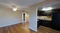 This single-floor residence offers a functional and cohesive layout with a warm, welcoming ambiance throughout. Rich hardwood flooring flows seamlessly across the main living area, adding character and warmth to the space. The living room is outfitted with a ceiling fan with integrated lighting, and neutral-toned walls provide a clean, versatile backdrop that complements a variety of interior styles. A hallway leads further into the unit, where a spacious walk-in closet can be spotted, offering ample storage solutions. The semi-open kitchen is positioned just off the main living area, separated by a half-wall that maintains a sense of openness while defining the space. The kitchen features dark espresso-toned cabinetry extending to the upper and lower sections, a gas range with black appliances, an overhead microwave, and a sink set beneath a well-lit countertop. Flush ceiling lighting brightens the kitchen area. White baseboards and trim throughout the unit provide a crisp contrast against the hardwood floors and neutral walls, contributing to a clean, well-maintained aesthetic overall.