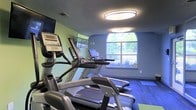 This fitness center offers a focused and functional workout environment designed to support an active lifestyle. The space is outfitted with a lineup of modern Matrix-brand cardio machines, including ellipticals and treadmills, each equipped with individual digital display consoles for tracking performance metrics. A wall-mounted television provides entertainment during workouts, adding to the overall comfort of the facility.

The room is painted in calming shades of green and blue, creating a refreshing and energizing atmosphere. Durable dark plank flooring runs throughout, providing both resilience and a clean, contemporary aesthetic. Overhead, large flush-mount oval light fixtures ensure bright, even illumination across the space.

A standout architectural feature is the arched window centered on the far wall, which floods the room with natural light and frames a view of lush greenery outdoors. This connection to the exterior enhances the ambiance, making workouts feel less confined and more invigorating. The textured ceiling adds a subtle design element that complements the overall interior finish. Compact yet thoughtfully arranged, this fitness center delivers an efficient and pleasant environment for cardiovascular exercise and general fitness routines.