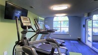 This fitness center offers a focused and functional workout environment designed to support an active lifestyle. The space is outfitted with a lineup of modern Matrix-brand cardio machines, including ellipticals and treadmills, each equipped with individual digital display consoles for tracking performance metrics. A wall-mounted television provides entertainment during workouts, adding to the overall comfort of the facility.

The room is painted in calming shades of green and blue, creating a refreshing and energizing atmosphere. Durable dark plank flooring runs throughout, providing both resilience and a clean, contemporary aesthetic. Overhead, large flush-mount oval light fixtures ensure bright, even illumination across the space.

A standout architectural feature is the arched window centered on the far wall, which floods the room with natural light and frames a view of lush greenery outdoors. This connection to the exterior enhances the ambiance, making workouts feel less confined and more invigorating. The textured ceiling adds a subtle design element that complements the overall interior finish. Compact yet thoughtfully arranged, this fitness center delivers an efficient and pleasant environment for cardiovascular exercise and general fitness routines.