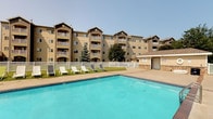 This multi-story residential complex is defined by its functional yet attractive exterior design, characterized by tan and brown siding, arched accent details, and pitched rooflines that give the building a structured, cohesive appearance. Each floor features private balconies, providing residents with personal outdoor space and views of the surrounding grounds.

The standout amenity of this property is the generously sized outdoor swimming pool, which takes center stage in the outdoor recreation area. The pool's clear turquoise water is complemented by a spacious concrete surround, accommodating multiple lounge chairs ideal for relaxing in the sun. A white perimeter fence encloses the pool area, providing both safety and a defined boundary between the pool and the surrounding lawn.

Adjacent to the pool, well-maintained grassy areas add greenery to the outdoor space, while mature evergreen trees contribute natural landscaping throughout the grounds. A small poolside utility or storage building with stone accents is also present, adding a practical element to the recreational area. The overall outdoor environment is open, bright, and well-suited for warm-weather leisure and community use.