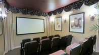 This private home theater is designed to deliver an authentic cinematic experience within a single, carefully appointed screening room. At the front of the room, a large projection screen dominates the wall, providing an expansive viewing surface ideal for films and presentations alike. Rows of dark upholstered theater-style seats are arranged in a tiered layout, ensuring clear sightlines from every position in the room. The seating is well-spaced with armrests, offering comfort during extended viewing sessions. The warm, golden-toned walls are illuminated by decorative wall sconces that cast a soft, ambient glow, contributing to the classic movie-house atmosphere. Ornate black-and-white swag valances line the upper perimeter of the room, adding a theatrical elegance that reinforces the cinema aesthetic. Draped curtain panels flank the screen area, further enhancing the formal presentation feel. Framed classic movie posters adorn the walls, completing the Hollywood-inspired décor. The deep red ceiling adds a dramatic, immersive quality to the space. Every design element in this room works cohesively to create an inviting, fully immersive private theater environment suited for entertaining guests or enjoying films in style.