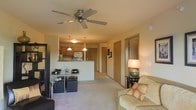 This single-floor residence is designed with an open-concept floor plan that seamlessly integrates the living, dining, and kitchen areas into one cohesive space. The living area is anchored by a ceiling fan with a pull-chain fixture, providing both air circulation and a functional lighting element. Plush carpeting throughout the main living space adds warmth and comfort underfoot.

The kitchen is positioned at the rear of the open layout and features wood-toned cabinetry, a white refrigerator, and a raised breakfast bar that serves as a natural divider between the kitchen and dining area. Two pendant lights hang above the bar, casting a warm glow over the space. The dining area accommodates a glass-top table paired with dark upholstered cube seating, offering a modern contrast to the warm surroundings.

A lamp-lit side table beside the sofa adds ambient lighting to the living area, complementing the overall warm tone of the space. A hallway visible in the background leads to additional rooms, with wood-toned doors lining the corridor. The layout is efficient and functional, making practical use of the available space throughout.