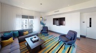 Located at 15400 NE 20th St in Bellevue, Washington, this contemporary amenity space offers a well-appointed, single-floor layout designed for both relaxation and social gatherings. Rich hardwood flooring flows throughout, unifying the open-concept interior and lending warmth to the clean, modern aesthetic defined by crisp white walls and recessed lighting.

The lounge area is anchored by a generously sized sectional sofa in deep blue tones, complemented by coordinating accent chairs and a bold striped area rug in teal, navy, and gold. Floor-to-ceiling windows flood the space with natural light and frame views of the surrounding community. A wall-mounted flat-screen television makes this area equally suited for casual viewing or social events.

Adjacent to the lounge, a dining area features a substantial wooden table with contemporary seating for multiple guests. A built-in kitchenette with wood-paneled cabinetry, stainless steel appliances including a microwave and beverage cooler, a sink, and a textured tile backsplash provides full functionality for entertaining. Striking mountain-themed artwork adorns the walls, adding an Pacific Northwest character to the interior.

Large sliding glass doors open directly onto an outdoor terrace, where a grill and additional seating invite al fresco gatherings with open-sky views — a seamless extension of the indoor space.