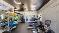 Located at 1361 Fox Run Dr. in Willoughby, Ohio, this well-appointed fitness center delivers a comprehensive workout environment within a smartly designed single-floor space. The room is finished with a striking dark ceiling adorned with sculptural circular pendant lights, creating a modern, high-energy atmosphere. Textured gray accent walls complement the crisp white trim and structural framing elements, lending the space a polished, contemporary aesthetic.

The flooring throughout is durable rubber gym matting, providing both safety and comfort during exercise. A row of cardio machines — including treadmills, ellipticals, and a recumbent bike — lines one side of the room, each positioned to face the glass-paned double doors that offer views to an adjacent area and allow natural light to filter in. On the opposite side, a tiered dumbbell rack provides a full range of free weights, while a dedicated functional training zone features a cable machine, an adjustable workout bench, and a well-stocked rack of stability balls, medicine balls, and foam rollers in various sizes. Full-length mirrors are strategically placed to expand the visual depth of the space and assist with form during exercise. The result is a compact yet fully equipped fitness facility that supports a wide range of training styles.
