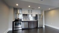 This well-appointed single-floor space showcases a thoughtfully designed interior with a consistent modern aesthetic throughout. The kitchen serves as a standout feature, boasting a full suite of stainless steel appliances including a range, dishwasher, and refrigerator. White upper cabinetry with frosted glass accents pairs elegantly with dark lower cabinets, creating a striking two-tone contrast. A shimmering mosaic tile backsplash adds texture and visual interest to the cooking area, while recessed lighting ensures the space is bright and functional. Light countertops complete the polished look of this culinary space.

Dark hardwood flooring runs throughout, grounding the interior with warmth and continuity. The hallway connects the various areas of the home with a clean, organized flow, featuring crisp white door trim and baseboards that contrast beautifully against the neutral gray walls. Louvered closet doors provide practical storage solutions along the corridor, while natural light filters in through windows visible in the adjoining rooms. The overall ambiance is one of contemporary elegance — understated, functional, and move-in ready, with a neutral palette that lends itself well to a variety of décor styles.