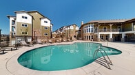 This expansive outdoor amenity space is designed to offer a resort-style experience within a residential community. At its centerpiece is a generously sized, freeform swimming pool with clear turquoise water, accessible via metal handrails and framed by a broad, tiled pool deck ideal for sunbathing and socializing. Adjacent to the pool is a bubbling hot tub with a decorative water feature, providing a relaxing retreat with scenic mountain views visible in the distance.

The surrounding deck is thoughtfully furnished with lounge chairs and seating areas, including a cozy fire pit lounge framed by natural stone landscaping. A wood-framed pergola offers shaded seating for those seeking relief from the sun, while wrought iron fencing encloses the entire pool area for privacy and safety.

The centerpiece building adjacent to the pool area is a striking two-story clubhouse structure, featuring stone pillar accents, warm wood-toned ceiling details, large panoramic windows, and an upper-level balcony with metal railings. The architecture blends natural stone masonry with contemporary siding elements, creating a warm and inviting aesthetic. Additional multi-story residential buildings surround the courtyard, contributing to an enclosed, community-centered atmosphere set beneath wide open blue skies.