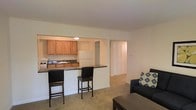 This single-floor space offers a functional and comfortable layout designed to maximize everyday living. The open-concept kitchen is equipped with warm wood-toned cabinetry, a white refrigerator, a sink, and ample counter space. A dark-topped breakfast bar with seating for two serves as a natural divider between the kitchen and the living area, making it an ideal spot for casual dining or entertaining. The living area features neutral wall-to-wall carpeting that contributes to a cozy ambiance, with enough room to accommodate a sofa and coffee table comfortably. Beyond the living space, a hallway leads to additional rooms, including a closeted space with louvered bifold doors that offer generous storage. The neutral color palette throughout — from the light beige walls to the carpeted floors — creates a clean, understated aesthetic that provides a versatile backdrop for a variety of interior styles. The open flow between the kitchen and living areas enhances the sense of space, making the layout feel connected and practical for everyday use.