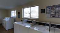This single-floor shared laundry facility is a functional and practical space designed to serve its users efficiently. The room is outfitted with a row of commercial-grade, coin-operated top-loading washing machines positioned along the main wall, providing ample capacity for laundry needs. The machines feature digital control panels, reflecting a modern and user-friendly design.

Natural light fills the room through large, multi-pane windows that overlook an adjacent residential building and surrounding greenery, creating a bright and open atmosphere. The walls are finished in a neutral, light tone that keeps the space feeling clean and welcoming.

Informational and instructional signage is prominently displayed throughout the room, including caution notices and usage guidelines posted near the machines, ensuring users are well-informed of proper operating procedures. A no-smoking sign is also clearly visible, indicating a strict policy for maintaining a clean environment.

The layout of the space is straightforward and accessible, with open floor space allowing easy movement around the equipment. Overall, this laundry facility is a well-maintained, no-frills communal space that prioritizes practicality, cleanliness, and convenience for its users.