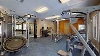 Located in Henderson, Nevada, this amenity space offers residents a comprehensive fitness and recreation experience. The fitness center is thoughtfully laid out across a single floor, featuring durable patterned carpet flooring and bright overhead fluorescent lighting that keeps the space well-illuminated. The gym is equipped with a wide variety of exercise options, including selectorized cable machines, a multi-station weight rack, a heavy punching bag with pull-up bar, a mini trampoline, a rowing machine, stationary bikes, and a full rack of dumbbells. Motivational words and phrases are painted directly onto the accent walls in warm orange and tan hues, adding an energizing atmosphere to the workout environment. Wall-mounted mirrors expand the sense of space, while a water station and informational kiosk provide added convenience. Beyond the fitness center, the outdoor area showcases a sparkling freeform pool with crystal-clear turquoise water, framed by mature towering palm trees and surrounded by lounge chairs perfect for relaxing in the Nevada sun. The pool area is set against the backdrop of a Mediterranean-style multi-story building, completing this well-rounded recreational retreat.