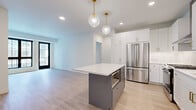 This thoughtfully designed single-floor unit offers a clean, contemporary aesthetic throughout its four rooms, blending modern finishes with functional layout. The open-concept kitchen is a standout feature, showcasing crisp white shaker cabinetry with matte black hardware, a white quartz peninsula, and classic subway tile backsplash. A full suite of stainless steel appliances — including a French door refrigerator, gas range with microwave, and dishwasher — completes the kitchen. Two globe pendant lights suspended from the ceiling add a stylish focal point above the peninsula. The living area flows naturally from the kitchen, bathed in natural light through large, black-framed grid windows that frame views of the surrounding streetscape. The bedroom continues the bright, airy theme with soft carpeting, a generous black-framed window, and clean white walls. The bathroom is elegantly finished with marble-style tile surround in the tub-shower combination, a white vanity with grey quartz countertop, and coordinating matte black fixtures throughout. Light wood-look flooring ties the main living spaces together, creating a cohesive and modern atmosphere across the entire unit.