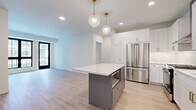 This thoughtfully designed single-floor unit offers a clean, contemporary aesthetic throughout its four rooms, blending modern finishes with functional layout. The open-concept kitchen is a standout feature, showcasing crisp white shaker cabinetry with matte black hardware, a white quartz peninsula, and classic subway tile backsplash. A full suite of stainless steel appliances — including a French door refrigerator, gas range with microwave, and dishwasher — completes the kitchen. Two globe pendant lights suspended from the ceiling add a stylish focal point above the peninsula. The living area flows naturally from the kitchen, bathed in natural light through large, black-framed grid windows that frame views of the surrounding streetscape. The bedroom continues the bright, airy theme with soft carpeting, a generous black-framed window, and clean white walls. The bathroom is elegantly finished with marble-style tile surround in the tub-shower combination, a white vanity with grey quartz countertop, and coordinating matte black fixtures throughout. Light wood-look flooring ties the main living spaces together, creating a cohesive and modern atmosphere across the entire unit.