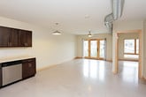 This single-floor unit showcases an industrial-modern aesthetic defined by polished concrete floors, exposed metallic ductwork running throughout, and clean neutral walls that create a bright, open atmosphere. The main living area is generously proportioned, featuring large wood-framed windows and glass-paneled French doors that invite abundant natural light and provide views of the surrounding neighborhood. A ceiling fan adds both function and a casual design element to the space.

The kitchen is well-appointed with dark espresso cabinetry offering ample storage, light-toned countertops, and a full suite of stainless steel appliances including a refrigerator, range, microwave, and dishwasher. The layout is efficient and thoughtfully organized, making it ideal for everyday cooking and entertaining.

The bathroom continues the modern industrial theme with a floating dark-toned vanity, an integrated white sink, stone-tiled shower surround, and a soaking tub. Exposed ductwork overhead reinforces the cohesive design language seen throughout the unit.

The doll-house view reveals a compact yet functional open floor plan where the kitchen, living area, and bathroom are logically arranged for ease of flow. Overall, the space combines urban industrial character with practical, contemporary finishes for a distinctive and comfortable environment.