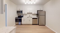 This single-floor space offers a practical and clean layout with a neutral color palette throughout. The living area is generously sized, featuring wall-to-wall carpet and a recessed entryway with a closet, providing a defined sense of arrival. Windows along the exterior walls allow natural light to brighten the carpeted rooms, contributing to an open and airy feel.

The kitchen is a functional highlight, outfitted with white shaker-style cabinetry extending to the ceiling for ample storage, light wood-look flooring, and a full suite of stainless steel appliances including a refrigerator, electric range, dishwasher, and microwave. A track lighting fixture overhead provides strong task illumination.

The bedrooms are comfortably scaled with plush carpeting, louvered bi-fold closet doors, and windows overlooking greenery. The bathroom is tastefully finished with classic white subway tile surrounding the soaking tub and shower, complemented by brushed gold fixtures. A separate vanity area features white cabinetry, a bright mirror, and light wood-look flooring, providing a clean and functional grooming space.