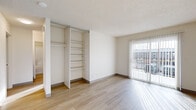 This single-floor unit presents a clean, neutral aesthetic throughout, with light-toned walls, textured ceilings, and a consistent use of light hardwood-style flooring across the main living areas. The open layout is anchored by a well-lit living space that benefits from a full sliding glass door fitted with vertical blinds, allowing natural light to stream across the floor and providing direct access to an outdoor area with exterior views.

The main living area connects to a kitchen space equipped with a refrigerator, light wood cabinetry, and hardwood-style flooring that continues seamlessly from the living area. An open-concept passage links the kitchen to the rest of the unit, aiding in overall flow and functionality.

Bedrooms feature soft carpeting and large windows dressed with vertical blinds that invite natural light. One bedroom includes built-in open shelving alongside a hanging rod, offering organized storage within a dedicated closet area. Additional carpeted rooms provide flexibility for various uses.

The bathroom features a full-size soaking tub with a shower combination and clean white surround with chrome fixtures. A dedicated utility closet houses a Rheem water heater and includes an overhead shelf with a hanging rod, maximizing the practicality of the space.