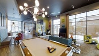 Located at 2101 Perennial Lane in Fort Collins, Colorado, this single-floor community amenity space offers a dynamic and energetic environment designed for both recreation and productivity. The interior showcases an eclectic, contemporary aesthetic defined by bold accent colors, vivid wall art, and a striking geometric chandelier that serves as a dramatic focal point overhead. Dark-painted ceilings contrast with bright, colorful walls, creating a lively and modern atmosphere throughout.

The space is anchored by a full-size billiards table positioned at the center of the main lounge area, surrounded by comfortable seating including a plush sofa and accent chairs. A flat-screen TV and media console provide additional entertainment options. Adjacent to the lounge, a dedicated workstation area features built-in desks and modern white chairs, ideal for residents seeking a productive workspace.

Large floor-to-ceiling windows and glass panel walls flood the interior with natural light while offering views of the surrounding exterior. Decorative curtains with a bold pattern add a touch of warmth and personality. The open layout connects seamlessly between zones, including what appears to be a dining or social area visible in the background, enhancing the communal flow of the space. The overall ambiance is vibrant, welcoming, and thoughtfully curated for an active community lifestyle.