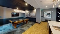 Located at 2000 South Blvd in Charlotte's South End neighborhood, this sophisticated single-floor amenity lounge is designed to offer a variety of social and recreational experiences within one expansive, open-concept space. The flooring throughout features a striking herringbone-pattern hardwood, providing a warm and cohesive visual foundation across all areas.

The main lounge area is anchored by a generously sized, tufted gray sectional sofa and a large caramel-toned leather channel-back sectional, both positioned to encourage conversation and relaxation. Large windows fitted with decorative drape panels allow ample natural light to fill the space. Pendant and floor lamps with sculptural bases add layered, ambient lighting throughout.

A bar area with teal upholstered counter seating sits beneath a dramatic cluster pendant light fixture, accompanied by a wall-mounted television, making it well-suited for social gatherings. A dedicated game lounge features a full-size pool table beneath a crystal chandelier, dark ceiling accents, and curated wall art, creating a moody, club-like atmosphere. A separate card table area and a built-in wine display rack round out the entertainment offerings, while bold artwork and eclectic décor elements lend the entire space a contemporary, design-forward character.