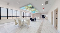 This contemporary single-floor commercial space is designed with a clean, upscale aesthetic that balances function and sophistication. Throughout the space, gleaming white marble-style porcelain flooring creates a bright, cohesive foundation, while expansive floor-to-ceiling windows along the perimeter flood the interiors with abundant natural light and offer sweeping panoramic views.

A standout feature of the space is its striking backlit ceiling panels, each displaying vibrant nature-inspired imagery including lush foliage and open skies, adding a distinctive artistic element to multiple rooms. The main gathering and dining area is generously proportioned, furnished with glass-top dining tables and modern chairs, making it well-suited for events or collaborative use.

The lounge area features multiple seating arrangements with leather sofas and chairs, creating a comfortable retreat alongside the panoramic windows. Two dedicated conference rooms are equipped with large multi-panel display screens, integrated A/V technology, and executive seating — ideal for meetings and presentations. The full kitchen is outfitted with stainless steel appliances, quartz countertops, a travertine-style backsplash, and ample cabinetry. The fitness center rounds out the amenities with modern cardio and strength equipment, rubberized flooring, and its own set of expansive windows.