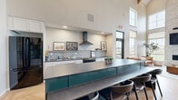 Located at 5532 38th St S in Fargo, North Dakota, this multi-functional community space seamlessly combines a fully equipped kitchen, a comfortable lounge, and an entertaining game room into one generously sized floor plan. The kitchen area is anchored by a striking teal island with dark stone countertops and bar seating, complemented by white shaker cabinetry, a patterned tile backsplash, a stainless range with wall-mounted hood, and black appliances. A sleek black steel-framed glass partition elegantly separates the kitchen from adjacent areas while maintaining an open feel. The lounge area is a standout, featuring soaring double-height ceilings, large floor-to-ceiling windows that flood the space with natural light and offer outdoor views, and a dramatic floor-to-ceiling stacked stone fireplace with a mounted flat-screen television. Warm herringbone hardwood flooring runs throughout the kitchen and lounge zones, adding a refined touch. The game room transitions to carpeted flooring and is lined with expansive windows, offering additional outdoor views. It includes a pool table beneath pendant lighting, high-top tables, dining seating, and vending machines, making it an ideal gathering space. Exposed wooden roof trusses visible from above add architectural character throughout.