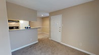 This single-floor residential space offers a straightforward and functional layout designed for comfortable everyday living. The main living area features wall-to-wall carpeting in a neutral beige tone, creating a warm and cohesive look throughout the space. Walls are painted in a soft, earthy beige, contributing to a calm and welcoming ambiance.

A half-wall bar counter separates the kitchen area from the living space, providing a convenient pass-through that maintains an open feel while defining each zone. The kitchen counter features a stone-like surface, and the kitchen area is illuminated by recessed lighting set into a slightly dropped ceiling section, adding a layered architectural detail to the space.

The entry area is anchored by a classic six-panel white door fitted with brass hardware, offering a traditional aesthetic accent against the neutral walls. Clean white baseboards run throughout, providing a crisp contrast to the carpeted floors and painted walls.

The overall design of this space prioritizes practicality and simplicity, with a neutral color palette that allows for versatile interior styling. Recessed lighting ensures adequate illumination, and the open connection between the kitchen and living areas supports a social and accessible flow throughout the unit.
