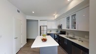 This single-floor residence showcases a thoughtfully designed contemporary interior with clean lines and a modern aesthetic throughout. The kitchen serves as the centerpiece of the space, featuring a striking two-tone cabinetry scheme that pairs crisp white upper cabinets with bold dark lower cabinets, creating a sophisticated contrast. A large central island with a white quartz countertop provides ample workspace and serves as a natural gathering point. The kitchen is equipped with stainless steel appliances, including a French door refrigerator and a built-in microwave, complemented by a black gas range and dishwasher. Glass-fronted upper cabinets add an open, airy feel, offering a glimpse of neatly arranged dishware. The undermount sink is paired with a modern faucet set against a clean backsplash. Recessed lighting illuminates the space evenly from above, highlighting the light-toned quartz countertops. Rich wood-look flooring runs throughout, grounding the space with warmth and texture. The open layout connects seamlessly to adjacent areas, enhancing the sense of flow and spaciousness across the residence.