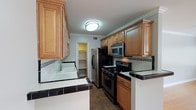 This single-floor space offers a clean, functional layout with thoughtful design details throughout. The main living area is bright and open, showcasing warm-toned hardwood flooring that runs seamlessly across the room. Crisp white crown molding and baseboards frame the light gray walls, lending a polished, cohesive look to the space. A ceiling-mounted light fixture provides overhead illumination, while a wall-mounted thermostat adds a practical touch near the entryway.

The kitchen is accessible via a peninsula-style pass-through featuring a dark-edged tile countertop, creating a natural visual boundary between the living and cooking areas. Inside, the kitchen is well-appointed with rich honey-toned wood cabinetry offering generous storage both above and below the counters. White tile countertops with dark trim accents complement the cabinetry and backsplash detail. Stainless steel appliances — including a gas range, over-the-range microwave, and refrigerator — provide a modern, practical cooking environment. A dedicated pantry or utility closet is accessible from within the kitchen, adding additional storage. Tile flooring in the kitchen provides a durable contrast to the hardwood in the main living area.