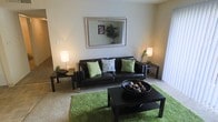 This single-floor residence offers a thoughtfully arranged living space designed for comfort and style. The living room serves as the heart of the home, featuring carpeted flooring accented by a vibrant green area rug that adds a pop of color and warmth to the room. A dark leather sofa, dressed with green and floral accent pillows, anchors the seating area and faces a sleek black coffee table holding a decorative bowl centerpiece. A framed rectangular mirror mounted on the neutral-toned wall reflects the room's natural greenery and ambient lighting, enhancing the sense of openness. Flanking the sofa are matching side tables, each topped with soft-glowing table lamps that contribute to the room's warm, inviting atmosphere. Tasteful plant accents and decorative branches add natural elements throughout the space. A large window allows natural light to filter in, brightening the interior and connecting the space to the outdoors. The overall design aesthetic balances clean lines with organic textures, creating a harmonious and livable environment that feels both functional and carefully curated.