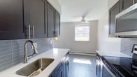 Located at 10900 Bustleton Avenue in the Northeast Philadelphia neighborhood of Somerton, this single-floor space offers a clean, modern aesthetic throughout. The standout feature is the beautifully updated kitchen, which showcases sleek dark gray shaker-style cabinetry that extends to the ceiling for generous storage. Crisp white quartz countertops provide a striking contrast, while a classic subway tile backsplash in a muted blue-gray tone adds texture and depth. The undermount stainless steel sink is paired with a contemporary arc faucet, and stainless steel appliances complete the kitchen's polished, cohesive look. Light tile flooring runs throughout, reflecting natural light that filters in through a window overlooking the surrounding neighborhood. A ceiling fan keeps the space comfortable year-round, and baseboard heating ensures warmth during cooler months. The open end of the kitchen flows into a dining or flex area, offering versatility in how the space can be arranged. The overall ambiance is bright, clean, and move-in ready, with thoughtful modern updates that make everyday living both functional and visually appealing.