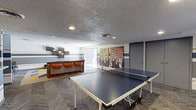The lounge and game room is a lively, multipurpose social space designed to encourage both relaxation and active recreation. The room's most prominent feature is a full-sized ping-pong table centered on the carpeted floor, providing ample space for gameplay and movement. A textured ceiling with recessed lighting keeps the atmosphere well-lit and energetic.

Along one wall, a built-in bar counter with a dark surface is paired with three orange upholstered bar stools, creating an inviting spot for casual conversation or refreshments. Floating shelves mounted above the bar add a stylish, functional touch to the area. The walls are accented with bold horizontal stripes in blue, yellow, and gray tones, contributing a dynamic, modern aesthetic throughout the space.

A large, colorful piece of wall art depicting a group of figures adds a creative focal point near the room's rear, complementing the overall playful ambiance. A glass door at the back allows natural light to filter into the space and suggests access to an adjacent area or exterior. The combination of game amenities, lounge seating, and eye-catching design elements makes this space well-suited for social gatherings and entertainment.