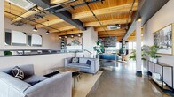 This thoughtfully designed single-floor space masterfully blends industrial and contemporary aesthetics, creating an inviting and functional environment throughout. The architectural character is defined by striking warm-toned wood plank ceilings supported by exposed structural beams, set against dark steel accents and black iron pipe conduit — a combination that lends the space a refined, urban loft quality.

Polished concrete floors run seamlessly throughout, reflecting natural light and contributing to the open, airy atmosphere. Expansive windows line the perimeter, flooding the interior with abundant daylight and offering views of the surrounding cityscape. Sheer dark curtains provide adjustable privacy without compromising the luminous quality of the space.

The open-plan layout is organized into several distinct zones. A communal lounge area features plush grey sofas and casual seating arrangements, ideal for relaxed conversation. A dedicated dining and collaboration area includes a large wood-top table surrounded by soft velvet chairs, positioned adjacent to a kitchenette with subway tile backsplash and pendant lighting. A stone-accented fireplace anchors one section, adding warmth and texture. Separate lounge nooks — one adorned with bold botanical wallpaper and a tufted leather sofa — offer intimate retreats within the broader open plan. Lush indoor plants are positioned throughout, softening the industrial materials and enlivening the overall ambiance.