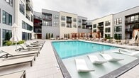 Located at 5505 W Northgate Rd in Rogers, Arkansas, this outdoor amenity space showcases a resort-style pool courtyard that serves as a central gathering destination. The space is defined by a generously sized, crystal-clear swimming pool with a clean geometric layout and in-pool lounge chairs positioned at the shallow entry, creating an inviting, resort-like atmosphere. Surrounding the pool is an expansive paver deck finished in light neutral tones, lined with rows of contemporary chaise lounges and patio seating arranged for both lounging and socializing. A series of elegantly appointed cabanas with warm wood-framed pergola structures and flowing curtains are positioned along the perimeter, offering shaded retreats. Market umbrellas and additional seating clusters are distributed throughout the deck, providing ample options for outdoor relaxation. The courtyard is framed on multiple sides by a modern multi-story building featuring large windows, private balconies, and a contemporary façade combining gray and cream tones with bold dark-framed fenestration. Neatly trimmed columnar evergreen trees and potted greenery add structured landscaping accents throughout the space, enhancing the polished, upscale ambiance of this well-appointed outdoor amenity area.
