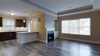 This single-floor space offers an open, flowing layout that seamlessly connects the living area to the kitchen. The living room is anchored by a classic white-mantled fireplace with a sleek black surround, flanked by built-in shelving and cabinetry that provide both storage and display space. A large window with white blinds allows natural light to fill the room, highlighting the warm, neutral-toned walls and rich dark hardwood-style flooring that runs throughout.

Crown molding and a tray ceiling detail add architectural refinement to the living area, lending an elevated, polished feel to the space. The open-concept kitchen is accessible via a raised breakfast bar with a granite-style countertop, creating a natural divide between the two areas while maintaining visual connectivity. Dark cabinetry in the kitchen contrasts with lighter countertops, and pendant lighting above the bar adds warmth and functional illumination. A window above the kitchen sink brings in additional natural light. The overall ambiance is clean, modern, and well-appointed, with thoughtful architectural details throughout.