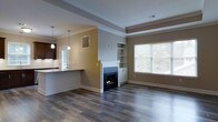 This single-floor space offers an open, flowing layout that seamlessly connects the living area to the kitchen. The living room is anchored by a classic white-mantled fireplace with a sleek black surround, flanked by built-in shelving and cabinetry that provide both storage and display space. A large window with white blinds allows natural light to fill the room, highlighting the warm, neutral-toned walls and rich dark hardwood-style flooring that runs throughout.

Crown molding and a tray ceiling detail add architectural refinement to the living area, lending an elevated, polished feel to the space. The open-concept kitchen is accessible via a raised breakfast bar with a granite-style countertop, creating a natural divide between the two areas while maintaining visual connectivity. Dark cabinetry in the kitchen contrasts with lighter countertops, and pendant lighting above the bar adds warmth and functional illumination. A window above the kitchen sink brings in additional natural light. The overall ambiance is clean, modern, and well-appointed, with thoughtful architectural details throughout.