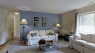 This single-floor residence offers a warm and inviting atmosphere, highlighted by thoughtful design choices and a cohesive color palette. The living room serves as the heart of the home, immediately drawing attention with its bold slate-blue accent wall, which provides a striking contrast against the surrounding neutral tones. Four framed black-and-white photographs are symmetrically arranged on the accent wall, adding a classic and curated aesthetic to the space. Soft, wall-to-wall carpeting in a warm beige anchors the room and contributes to its comfortable ambiance. A plush white slipcovered sofa, adorned with chevron-patterned throw pillows in coordinating blues and grays, serves as the focal seating piece, complemented by a matching white armchair. An ornate wrought-iron coffee table with a lower shelf sits at the center, offering both style and function. Ambient lighting is provided by elegant floor lamps with classic cream shades, flanking the seating arrangement and casting a soft, welcoming glow. Decorative accents, including potted greenery and a sculptural branch arrangement, bring natural elements into the interior. The smooth white ceiling enhances the sense of openness, making the living area feel airy and well-proportioned throughout.