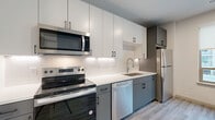 This thoughtfully designed one-bedroom, one-bathroom unit offers a modern and functional living experience across a single floor. Light wood-tone luxury vinyl plank flooring runs throughout the space, lending warmth and visual continuity between rooms.

The kitchen is a standout feature, boasting a clean two-tone design with crisp white upper cabinetry and slate-gray lower cabinets, all accented by matte black hardware. A white subway tile backsplash, light quartz countertops, and a full suite of stainless steel appliances — including a range, microwave, dishwasher, and refrigerator — complete this well-appointed cooking space. Recessed lighting illuminates the area effectively.

The open living area benefits from a large picture window fitted with roller shades, allowing for natural light while maintaining privacy. A stylish sliding barn door connects key areas of the unit, adding a modern architectural detail and maximizing usable floor space.

The bedroom includes access to a walk-in closet with built-in shelving, offering practical storage solutions. The bathroom is clean and contemporary, featuring a glass-enclosed walk-in shower, a gray vanity with white countertop, and matte black fixtures and accessories that tie cohesively into the unit's overall design language. Recessed lighting and wood-look flooring carry through here as well, maintaining a unified aesthetic.