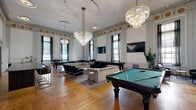 Located at 101 W 10th St in Wilmington, Delaware, this expansive single-floor lounge is a masterclass in neoclassical elegance blended with modern leisure. The space commands immediate attention with soaring ceilings adorned with gilded medallion friezes and intricate crown molding, all set against crisp white walls that amplify the sense of grandeur. Underfoot, warm-toned herringbone hardwood flooring adds a timeless sophistication throughout.

Towering floor-to-ceiling windows dressed with dark curtains flood the interior with natural light while offering a refined aesthetic. Cascading crystal chandeliers anchor the room visually, providing dramatic illumination that complements the ornate architectural detailing above.

The space is thoughtfully arranged to accommodate a variety of social activities. Two full-size billiard tables serve as centerpieces, while a dedicated poker table with black-and-gold upholstered seating offers a sophisticated gaming corner. Multiple lounge seating arrangements, featuring plush velvet sofas and leather armchairs, are positioned throughout for relaxed conversation. A stylish bar counter with gold-accented bar stools and wall-mounted flat-screen televisions further enhance the entertainment experience.

Decorative potted tropical plants are strategically placed to introduce organic texture and life. The ornate gilded entrance doorway provides a stately arrival moment, completing this remarkable space.