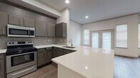 This single-floor residence offers a thoughtfully designed layout that balances functionality and modern style. The open-concept kitchen is a standout feature, showcasing gray cabinetry, a mosaic tile backsplash, gleaming quartz countertops, a large peninsula with an integrated sink, and a full suite of stainless steel appliances including a refrigerator, electric range, and microwave. The kitchen flows seamlessly into the living area, which is bathed in natural light from large windows and a set of glass French doors. A corner fireplace with a classic white mantle serves as the focal point of the living space, complemented by neutral walls and wood-look flooring that runs throughout the home. The bedrooms are generously sized, with at least one featuring a spacious walk-in closet equipped with wire shelving for ample storage. The bathrooms are finished with clean white tile surrounds accented by a distinctive checkered gray-and-white tile border, paired with a soaking tub and shower combo. Consistent wood-look flooring ties the spaces together, while recessed lighting and crown molding add polished finishing touches throughout this well-designed residence.