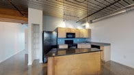 Artek Lofts showcases a thoughtfully designed single-floor space that blends industrial character with modern comfort. Polished aggregate concrete floors run continuously throughout, reflecting natural light and lending a clean, open feel to every room. The ceilings feature a striking mix of corrugated metal panels and warm wood sections, with exposed silver ductwork reinforcing the loft's industrial aesthetic.

The kitchen serves as a central hub, anchored by a large island with dark granite countertops and natural wood cabinetry. Black appliances, including a refrigerator, range, microwave, and dishwasher, provide a bold contrast against the light maple cabinets and teal backsplash. Pendant lights hang from a track-lighting system overhead, adding both function and style.

The living areas are bright and versatile, lined with rows of windows fitted with horizontal blinds that invite generous natural light while offering views of surrounding greenery. These open rooms offer flexible use of space beneath their distinctive ceiling treatments.

A clean, well-appointed bathroom features white subway tile wainscoting, large-format floor tile, a pedestal sink, and a tub-shower combination. Hallways are wide and bright, with polished floors continuing seamlessly throughout. A walk-in storage area and glass-panel entry door complete this well-equipped, architecturally distinctive loft space.
