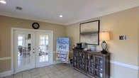 This single-floor community space makes an immediate impression with its warm and inviting reception area. The entry is finished with neutral beige walls, crown molding, and light tile flooring that lends a clean, classic aesthetic throughout. Recessed lighting complements the natural light that filters in through elegant white-framed French doors, which open to an adjoining area and offer a bright, airy feel to the space.

A rustic dark wood cabinet with glass-paneled doors serves as a functional and decorative focal point, topped with a table lamp and a coffee station for the convenience of residents and guests. Above it, a large framed landscape artwork adds a refined touch to the wall. A round wall clock anchors the opposite side of the room, adding to the homey atmosphere.

The space is clearly oriented around community and hospitality, as reflected by prominent resident referral signage near the entrance. Crown molding details and recessed lighting throughout contribute to a polished, well-maintained environment. Overall, the space balances practicality with a welcoming residential charm, making it a comfortable and functional hub for community interaction.