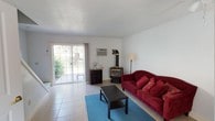 This two-floor home offers a clean, straightforward layout with practical features throughout. The living room serves as the heart of the main level, finished with light tile flooring and anchored by a classic freestanding gas stove with black piping — a standout functional element that adds character to the otherwise neutral space. A sliding glass door floods the room with natural light and provides direct access to the outdoor area, where greenery is visible beyond. A wall-mounted AC unit ensures year-round comfort. The living area connects seamlessly to the kitchen and dining space, visible through an open pass-through. The kitchen is well-equipped with white cabinetry, tile countertops, white appliances including a range, dishwasher, and microwave, and a large window above the sink that brings in additional natural light. The tile backsplash complements the overall bright, clean aesthetic. Upstairs, the bedrooms feature neutral carpet flooring, soft grey walls, and ceiling fans for ventilation. Each room receives natural light through curtained windows with views of surrounding trees. The rooms offer a calm, understated atmosphere with simple, functional finishes throughout.