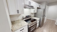 The Hamilton 2B Premium apartment offers 930 square feet of thoughtfully designed single-floor living space, featuring 2 bedrooms, 2 bathrooms, and 2 closets. The open-concept kitchen and living area serves as the heart of the unit, with light wood-look flooring, recessed lighting, and a textured ceiling. The kitchen is equipped with white shaker-style cabinetry with black hardware, granite countertops, a peninsula with a sink, and stainless steel appliances including a refrigerator, range, and dishwasher. The spacious living room offers large windows with blinds and a private exterior door, allowing ample natural light. Both bedrooms feature soft wall-to-wall carpeting and neutral gray tones. The primary bedroom, at 150 square feet, includes access to a dedicated closet and an en-suite bathroom. The secondary bedroom benefits from a large window with greenery views. Both bathrooms are finished with granite-topped vanities, wood-look flooring, and tub-shower combinations with tile surrounds. Two closets provide generous storage throughout the unit. The overall aesthetic is clean and contemporary, with a consistent neutral color palette, white trim, and modern fixtures creating a bright, cohesive living environment.