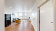 This thoughtfully laid-out 1-bedroom, 1-bathroom unit offers a clean, modern aesthetic across a single floor. The open-concept living and kitchen area serves as the heart of the home, with warm hardwood-style flooring running throughout and crisp white walls that enhance the bright, airy atmosphere. The living room is generously proportioned, featuring multiple windows that flood the space with natural light and offer views of the surrounding neighborhood. The kitchen is well-equipped with dark espresso cabinetry, light countertops, recessed lighting, and black appliances including a refrigerator, dishwasher, and range — providing an efficient and functional cooking space that flows seamlessly into the living area. The bedroom is a comfortable, sunlit retreat with a large window framing outdoor views and ample space for furnishings. The full bathroom features hardwood-style flooring, a wood-toned vanity with a white countertop, and a tub-shower combination. A dedicated in-unit laundry room with a stacked Whirlpool washer and dryer and a built-in shelving rod adds a practical and convenient touch. The overall design is clean and contemporary, making efficient use of every square foot.