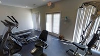 This single-floor fitness facility is designed to support a well-rounded workout routine. The space is outfitted with a selection of cardio and strength training equipment, including a stationary bike, a treadmill, and additional resistance machines, providing users with a variety of exercise options. The flooring consists of durable dark rubber matting, a practical choice for a high-use workout environment that offers both cushioning and traction. Large windows fitted with horizontal blinds line the walls, allowing generous amounts of natural light to fill the room while still offering privacy control. Vertical blinds on an adjacent window further contribute to light management throughout the day. The walls are finished in a neutral gray tone, creating a clean and focused atmosphere. A wall-mounted storage rack provides convenient organization for accessories and supplies. An exit sign and ceiling ventilation are visible overhead, reflecting the room's attention to safety and air circulation. The overall ambiance is functional and bright, offering a comfortable and practical environment for fitness activity.