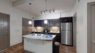 This single-floor unit offers a well-designed layout that balances modern aesthetics with functional living. The kitchen serves as a standout feature, showcasing dark espresso cabinetry paired with a mosaic tile backsplash, granite countertops, and a full suite of stainless steel appliances, including a refrigerator, range, and built-in microwave. A curved kitchen island with an undermount sink provides additional prep space and casual seating, while pendant lighting and a track light fixture illuminate the area with a contemporary flair. Rich wood-look flooring runs throughout the main living areas, adding warmth and visual continuity to the space.

The living area is open and versatile, featuring a ceiling fan and neutral white walls that create a clean, adaptable backdrop. The bedroom connects directly to the bathroom, which features matching dark cabinetry, a granite-topped vanity, a framed mirror, and wood-look flooring. A walk-in closet adjacent to the bathroom provides ample storage with wire shelving systems. The overall design is cohesive and modern, with consistent finishes and fixtures throughout that contribute to a polished, move-in-ready atmosphere.