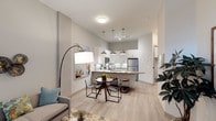 This thoughtfully designed single-floor residence offers a seamless open-concept layout that connects the living, dining, and kitchen areas into one cohesive, functional space. The kitchen is a standout feature, outfitted with stainless steel appliances, white shaker cabinetry, a subway tile backsplash, and a breakfast bar peninsula with pendant lighting that adds a warm, modern ambiance. Light wood-style flooring runs throughout, creating a clean and airy feel that ties the entire space together. The dining area sits adjacent to the kitchen, comfortably accommodating a round table with modern chairs — an ideal setting for casual meals or entertaining. The living area is well-proportioned, featuring neutral-toned walls that allow for versatile décor choices. Concrete accent walls lend an industrial yet refined character to the space, balancing the softness of the light palette. A full-length arc floor lamp adds a stylish focal point to the living zone. The layout is efficient and welcoming, with natural sightlines across the entire open floor plan. Overall, the space strikes a balance between modern urban design and everyday livability, making it a comfortable and visually appealing environment.