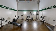 Located at 31 Pope Park Hwy in Hartford, Connecticut, this fitness room offers a well-equipped exercise space with an industrial-inspired aesthetic. The room features soaring ceilings supported by exposed concrete columns, giving the space an open, airy feel despite its compact footprint. Polished concrete floors run throughout, adding a durable and clean finish suited to an active environment.

The walls are painted white and accented with a bold green horizontal stripe, complemented by motivational text decals reading "STRENGTH," "MOTIVATION," and "FITNESS," creating an energizing atmosphere. A wall-mounted television provides entertainment during workouts.

The room is outfitted with a diverse selection of equipment, including a treadmill, elliptical trainer, stationary bikes, an inversion table, a Total Gym cable machine, battle ropes, a pull-up and dip station, exercise mats, and mirrors for form-checking. This variety supports both cardio and strength training routines.

A sleek glass partition wall with black-framed panels and a glass door separates the main exercise area from an adjacent space, allowing natural light to flow through while maintaining a modern, open visual connection. The overall ambiance is functional and motivating, making it a practical fitness facility for residents or building occupants.